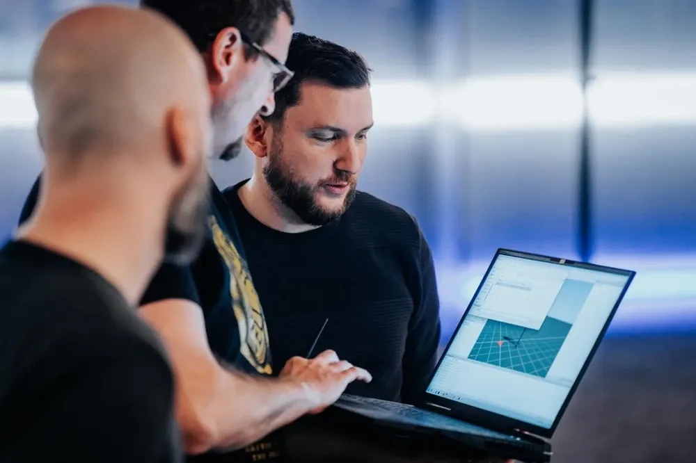 Three developers gathered around a laptop, reviewing work in a 3D viewport